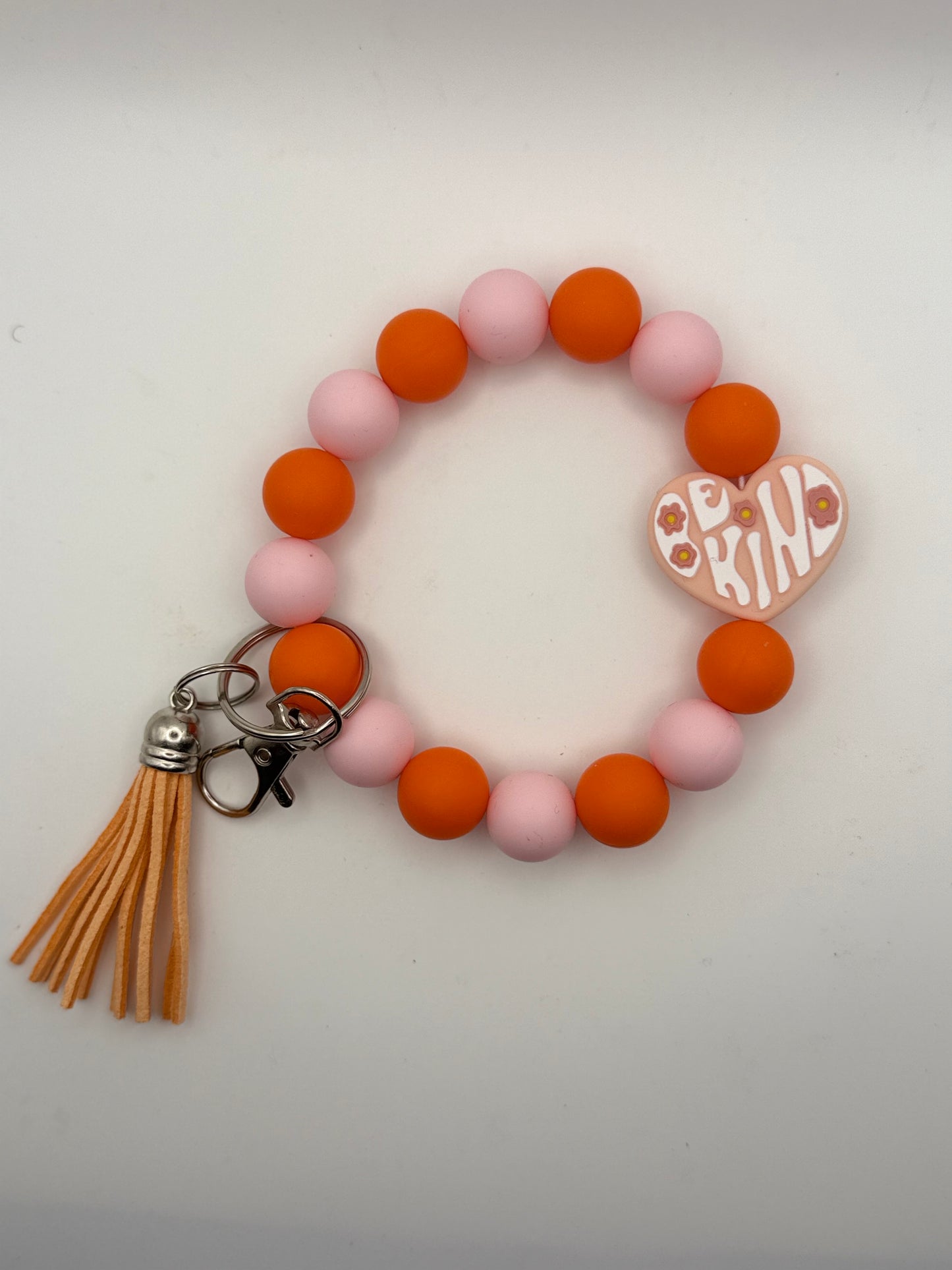 Bracelet with pink and orange beads, tassel, and 'Be Kind' charm on a white background