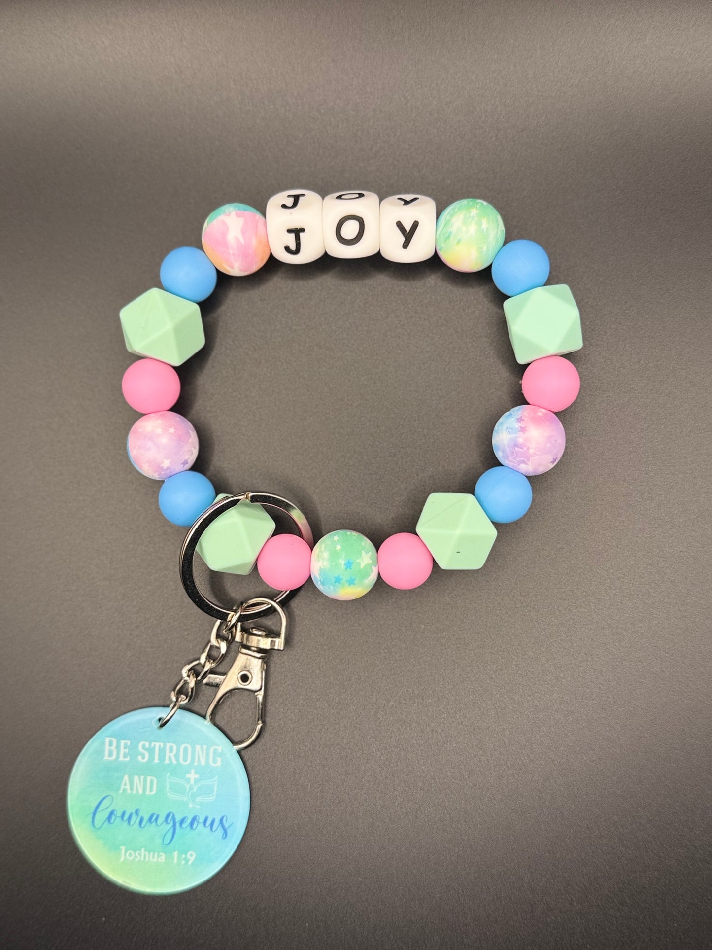 Colorful beaded bracelet with a keychain and with letter beads spelling 'JOY' on a gray background