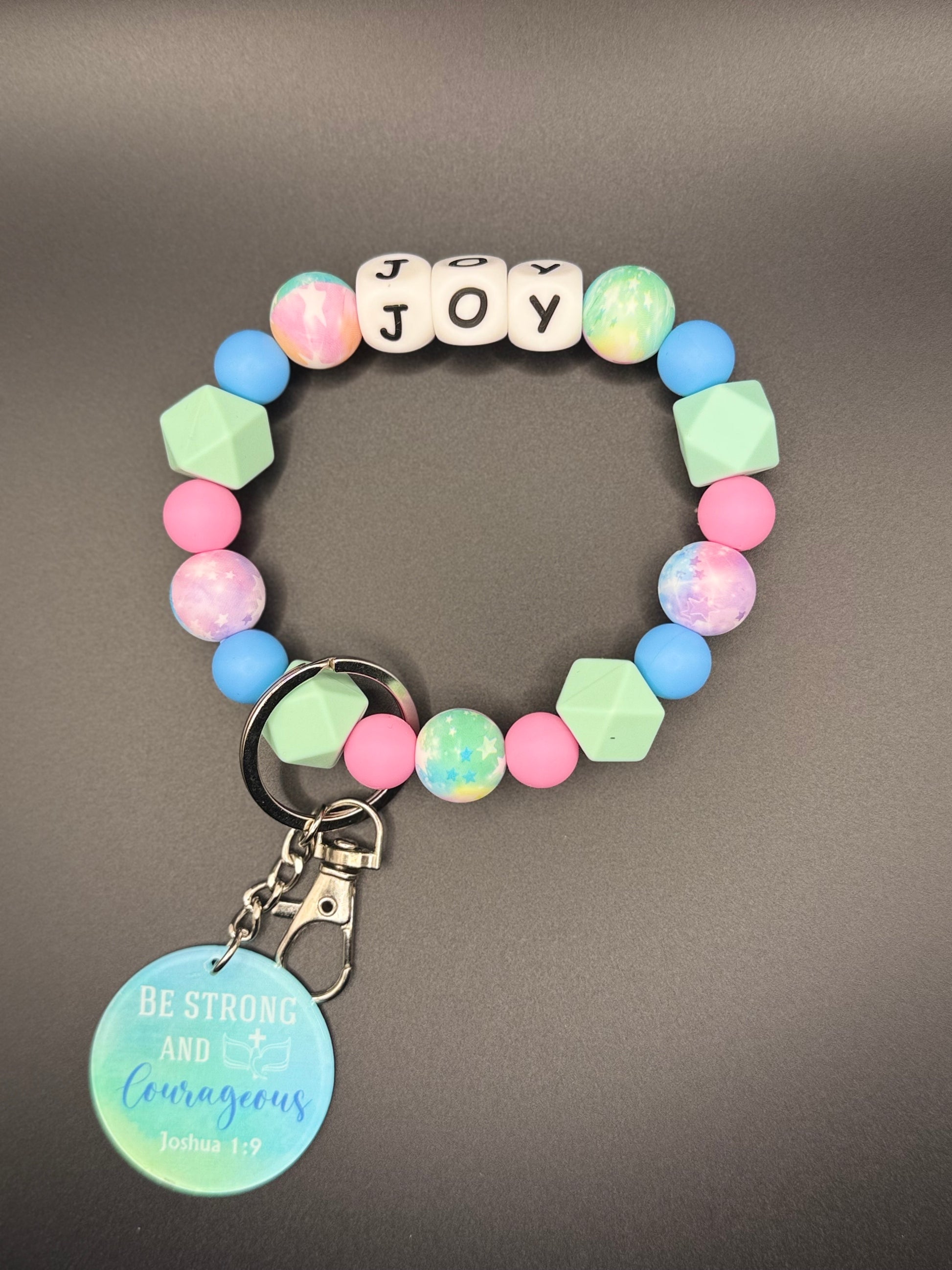 Colorful beaded bracelet with a keychain and with letter beads spelling 'JOY' on a gray background