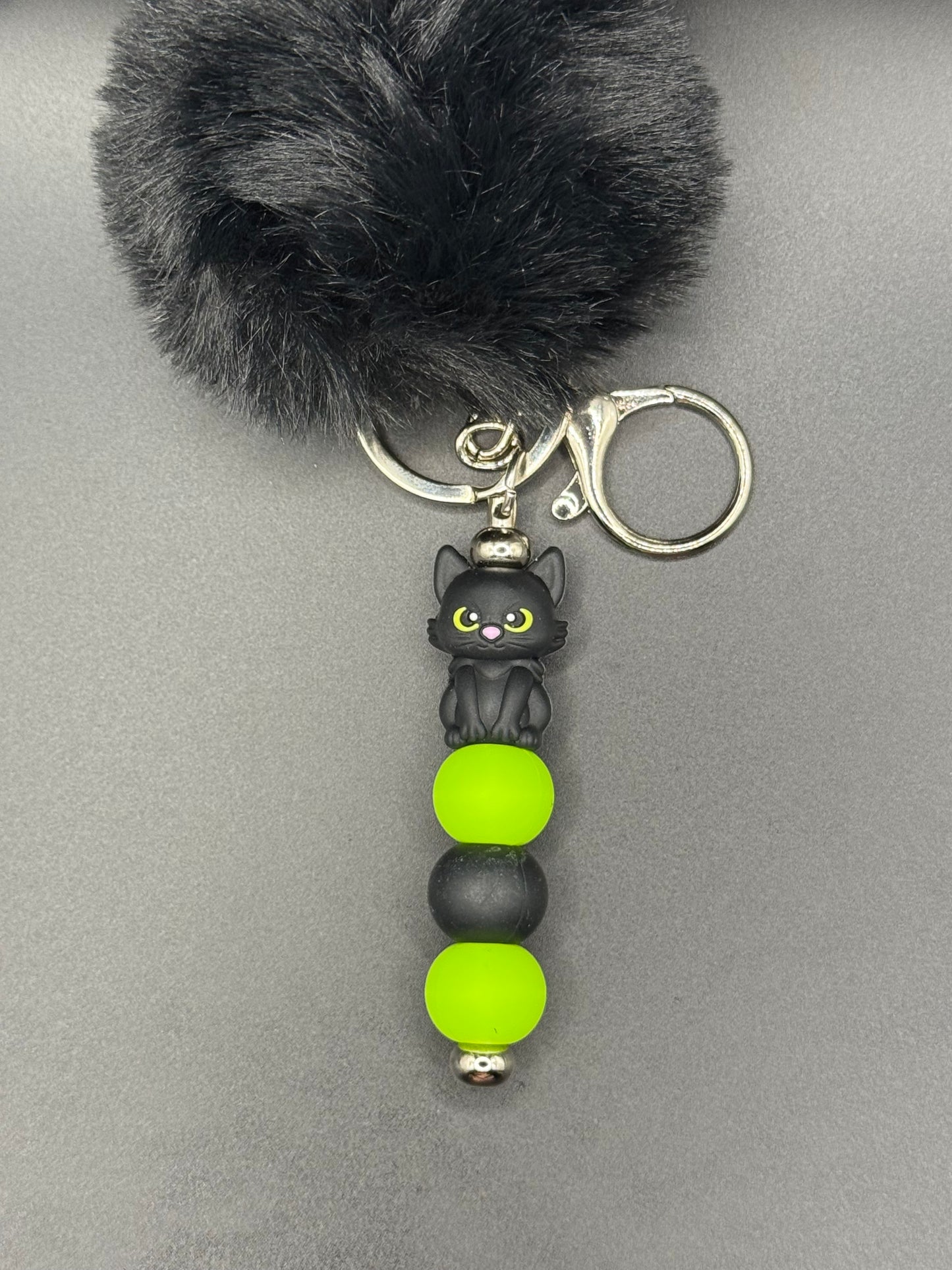 Keychain with black pom-pom, cat charm, and green beads on a gray background