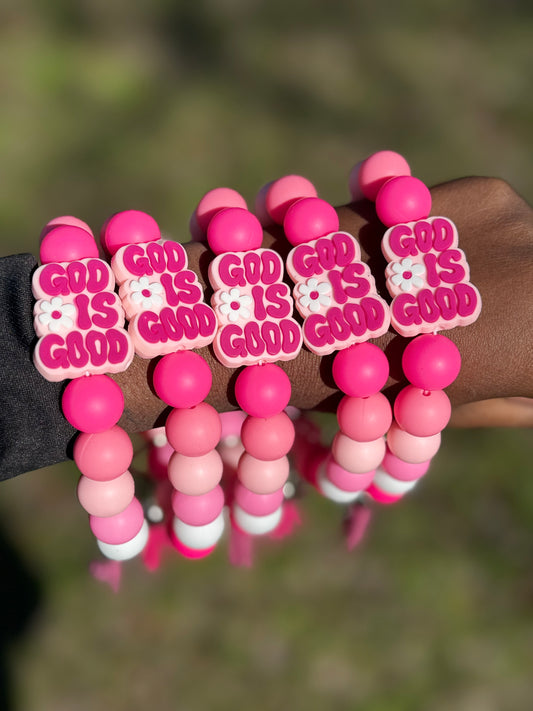 Pink beaded bracelet with 'GOD IS GOOD' text on a wrist against a blurred green background